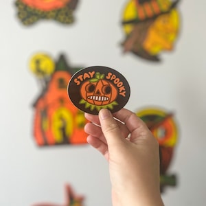 May include: A round sticker with a brown background and the text "Stay Spooky" in white. A cartoon pumpkin with a scary face is in the center of the sticker.
