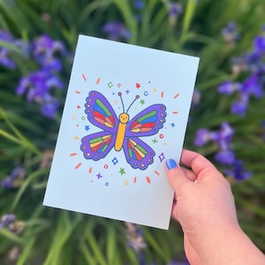 May include: A light blue card featuring a colorful cartoon butterfly with a smiling face. The butterfly has purple, red, yellow, green, and blue wings. Surrounding the butterfly are colorful stars, circles, and confetti. The card is held in front of a blurred background of purple flowers.