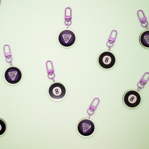 May include: Eight purple and black keychains with a purple eight ball design and a purple triangle with the text "What if it all works out?" on the other side.