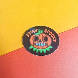 May include: A round sticker with a black background and a cartoon pumpkin with a toothy grin. The text "Stay Spooky" is written in white around the pumpkin.