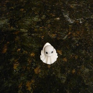 May include: A small, white, ghost-shaped figurine with a rounded head, a ruffled base, and two dark, circular eyes. The figure has a smooth, matte texture and is positioned upright, displaying a simple, stylized form with a pointed top.