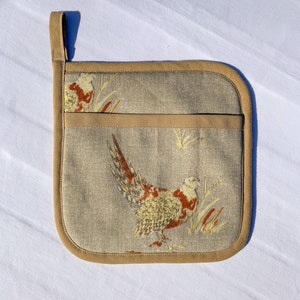 May include: A beige pot holder with a brown trim and a pheasant design. The pheasant is brown, yellow, and orange.