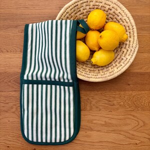 May include: A double oven glove featuring a white and dark green vertical striped pattern with solid dark green trim and a hanging loop, positioned next to a round woven basket filled with six bright yellow lemons on a wooden surface.