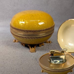 Op de afbeelding: Een ronde, gele, getextureerde doos met deksel, een goudkleurige metalen rand en decoratieve voetjes. Daarnaast onthult een bijpassende open houder een goudkleurig metalen aanstekermechanisme in de basis, met een gladde, crèmekleurige binnenkant.