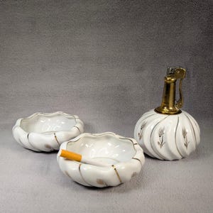 May include: A white ceramic cigarette lighter and two matching ashtrays with gold accents and a wheat design. The lighter has a brass top and a flip-top lid.
