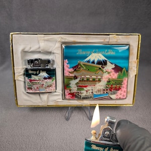 May include: A vintage souvenir set featuring a lighter and a decorative tile, both adorned with a Japanese landscape design. The lighter is teal with a detailed image of a pagoda and Mount Fuji. The tile has a similar design with the text "Memory of Japan & Kona".