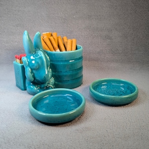 Peut inclure: Un porte-cigarettes et allumettes en céramique turquoise avec un motif de lapin. Le porte-cigarettes a une section cylindrique pour les cigarettes et une section rectangulaire pour les allumettes. Il y a deux petits cendriers en céramique turquoise.