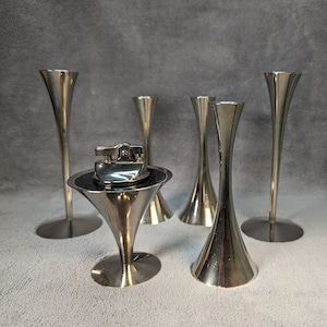 May include: Five silver metal candle holders with a silver metal lighter in the center. The candle holders are all different shapes and sizes.