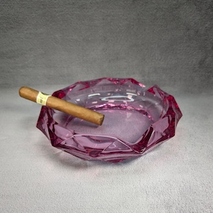 Könnte beinhalten: Ein rosa Aschenbecher aus Glas mit facettiertem Design. Eine braune Zigarre liegt am Rand des Aschenbechers.
