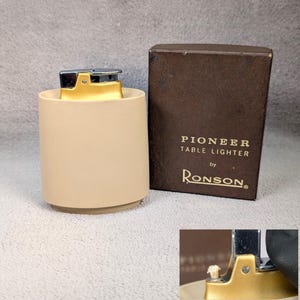 May include: A vintage Ronson Pioneer table lighter in a beige cylindrical holder. The lighter has a gold-colored metal top and a silver-colored ignition mechanism. The original box is brown with the text "PIONEER TABLE LIGHTER by Ronson."