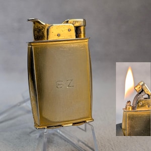 May include: A gold-toned lighter with a rectangular body and a hinged lid. The lighter has a small, engraved inscription that reads "GZ". The lighter is shown with the lid open and a flame coming from the top.