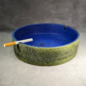 Peut inclure: Un cendrier en céramique bleu et vert avec une cigarette posée sur le bord. Le cendrier a un extérieur rugueux et texturé et un intérieur lisse et bleu.