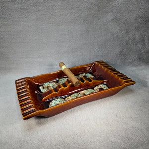 May include: A brown ceramic ashtray with a decorative design and a single cigar resting in the groove.