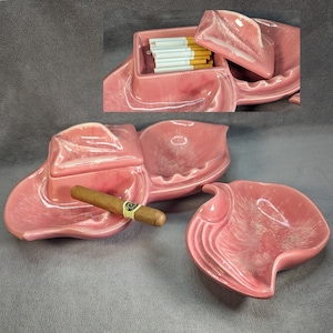 May include: A pink ceramic ashtray with three compartments. The largest compartment is shaped like a leaf and has a cigar resting in it. The middle compartment is a round bowl. The smallest compartment is a rectangular box with a lid that is open and has cigarettes inside.