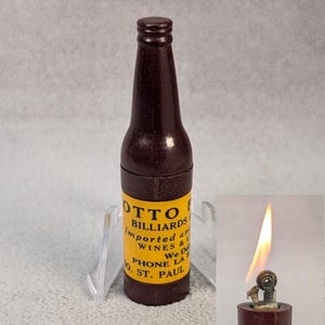 May include: A dark brown, bottle-shaped lighter with a yellow label featuring the text "OTTO BILLIARDS Imported Wines & We De Phone LA 10 St. Paul". The lighter is lit, showing a flame.