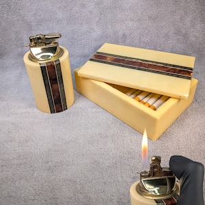 Pode incluir: Um conjunto vintage de isqueiro e caixa de cigarros cor marfim. O isqueiro tem um topo dourado e um design de listras verticais em marrom e preto. A caixa de cigarros está aberta, revelando cigarros dentro. O isqueiro está aceso, mostrando uma chama.