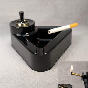 May include: A black, triangular ashtray with a built-in lighter. The ashtray has a small, circular platform with a gold-colored ring and a black knob. A lit cigarette rests in the ashtray. The lighter is shown lit in the bottom right corner.