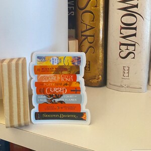 May include: A die-cut sticker featuring a vertical stack of seven colorful book spines with visible titles including The Priory of the Orange Tree, Harry Potter, City of Bones, The Poppy War, Cursed, A Court of Silver Flames, and Firekeeper's Daughter.