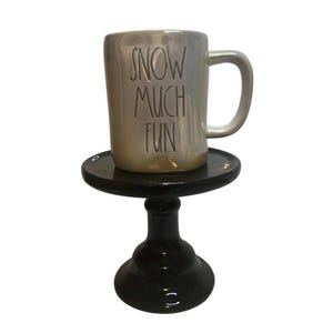 May include: A pearlescent ceramic mug with the words "SNOW MUCH FUN" in black lettering. The mug is on a small, black, pedestal-style cake stand. The stand has a round top and a decorative base.
