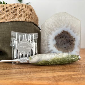 May include: A crochet hook with a marbled green and white resin handle, a clear plastic bag containing several silver metal hook tips with a circular label reading THE RESIN HOOK SHED, and a large translucent white geode with a dark centre.
