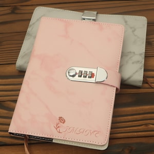 May include: Pink marble-patterned journal with a combination lock. The journal has a floral design and the word "IMANE" printed on the cover. A gray marble-patterned journal is visible in the background.