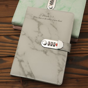 Può includere: Un diario con motivo a marmo bianco con una serratura a combinazione argentata e un cinturino argentato. Il diario ha il testo "Proverbi 3:3 Scrivili sulla tavoletta del tuo cuore" stampato sulla copertina.