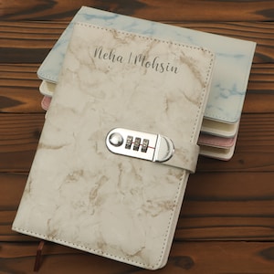 May include: A stack of personalized marble-patterned notebooks. The top notebook has a combination lock and the names "Neha | Mohsin" printed on the cover. Other notebooks in the stack are light blue, pink, and white.