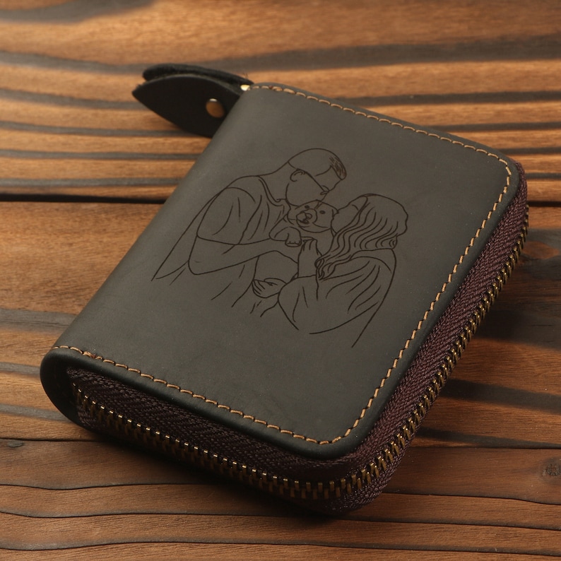 May include: A black leather zippered wallet with a personalized engraved design of a couple and their pet. The wallet has brown stitching and a gold zipper.