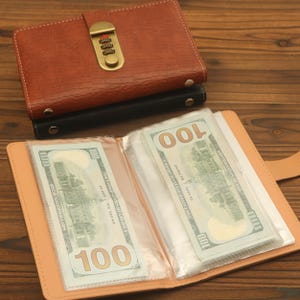 Op de afbeelding: Een beige lederachtige portemonnee met doorzichtige plastic hoesjes met twee briefjes van 100 dollar. Daarboven een gesloten bruine portemonnee met een gestructureerd oppervlak en een messingkleurig cijferslotmechanisme.