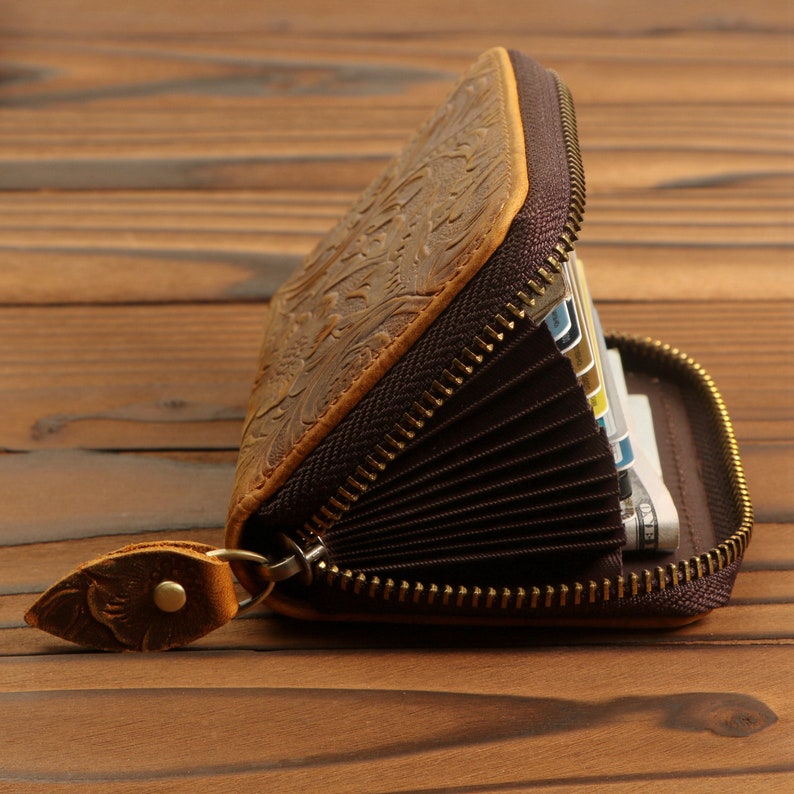 May include: A brown leather wallet with a zipper closure and intricate floral embossed design. The wallet is open to reveal multiple card slots and a bill compartment.