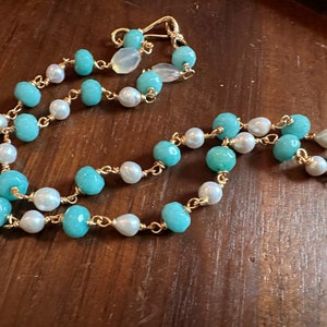May include: A delicate necklace featuring alternating turquoise and white pearl beads strung on a gold-colored chain. The necklace has a simple clasp and is displayed on a wooden surface. The jewelry is a necklace.
