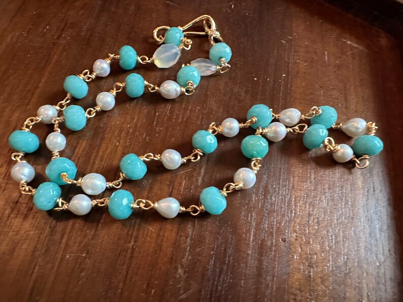 May include: A delicate necklace featuring alternating turquoise and white pearl beads strung on a gold-colored chain. The necklace has a simple clasp and is displayed on a wooden surface. The jewelry is a necklace.