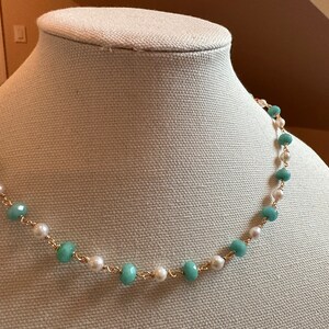 May include: A delicate necklace featuring alternating light blue and white beads strung on a gold-colored chain. The necklace is displayed on a mannequin, showcasing its design and color contrast. The beads are round and smooth, creating a classic jewelry piece.