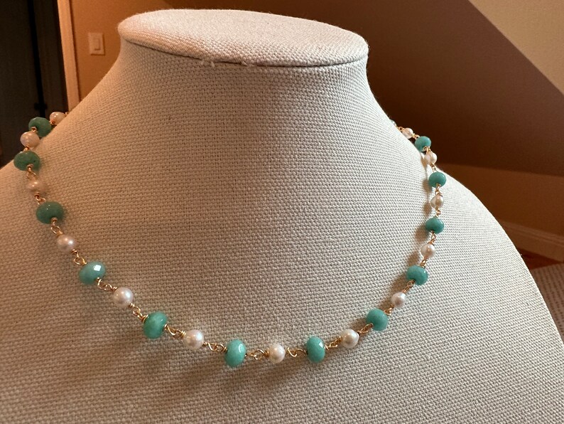 May include: A delicate necklace featuring alternating light blue and white beads strung on a gold-colored chain. The necklace is displayed on a mannequin, showcasing its design and color contrast. The beads are round and smooth, creating a classic jewelry piece.