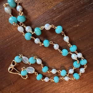 May include: A delicate necklace featuring alternating turquoise and white beads strung on a gold chain. The necklace has a lobster clasp closure. The beads are a mix of round and irregular shapes, creating a unique and elegant design.