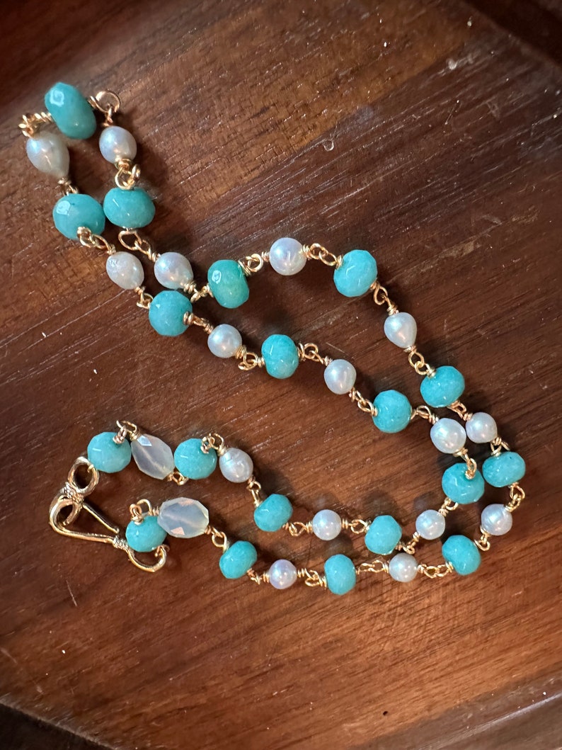 May include: A delicate necklace featuring alternating turquoise and white beads strung on a gold chain. The necklace has a lobster clasp closure. The beads are a mix of round and irregular shapes, creating a unique and elegant design.