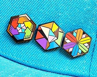May include: Three colorful geometric enamel pins with black outlines. The pins are in shades of orange, yellow, purple, and blue. The pins are on a blue fabric background.
