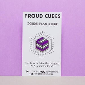 May include: A white card with the text "PROUD CUBES" and "PRIDE FLAG CUBE" in black. A purple, white, and grey geometric cube is shown in the centre of the card. The text "Your Favourite Pride Flag Designed As A Geometric Cube!" is below the cube. The text "@proud.zebra @proudzebra www.proudzebra.com" is at the bottom of the card.