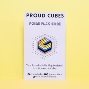 May include: A white card with the text "PROUD CUBES" and "PRIDE FLAG CUBE" in black. A yellow, blue, and white geometric cube pin is shown on the card. The text "Your Favorite Pride Flag Designed As A Geometric Cube!" is also on the card. The card has the social media handles "@proud_zebra" and "@proudzebra" and the website "www.proudzebra.com".