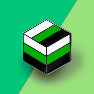 May include: A black, white, and green enamel pin shaped like a cube. The pin has three horizontal stripes, with the top stripe being white, the middle stripe being green, and the bottom stripe being black.