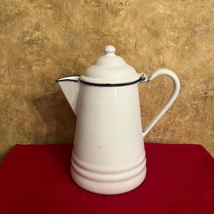 May include: A white enamel coffee pot with a lid and handle. The pot has a black trim around the spout and lid. The pot is sitting on a red surface.