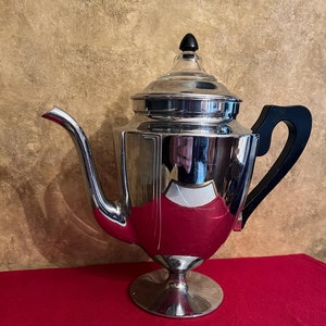 May include: A silver and black coffee pot with a glass lid and a black handle. The pot is sitting on a red surface.