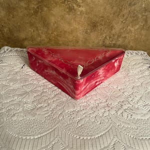 May include: A triangular, red and white marbled box with a clear lid. The box has a decorative design along the top edge. The box is sitting on a white, lace tablecloth.