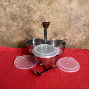 May include: A stainless steel condiment set with a wooden handle, two small bowls, and a central container with a clear lid. Includes two clear lids and a spoon. The set is on a red surface.