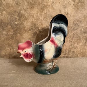 May include: A ceramic rooster-shaped planter or decorative vessel featuring a glossy finish. The rooster is painted with a cream-colored body, a vibrant red comb and wattle, and dark green and black accents on the neck, wings, and tail feathers.