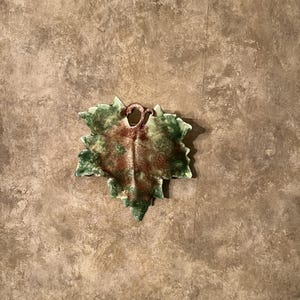 May include: A ceramic leaf-shaped dish with a green and brown color scheme. The dish has a textured surface and a small loop for hanging. The leaf design is detailed, with visible veins and edges. The dish is likely for decorative purposes.