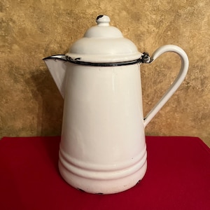 May include: A white enamel coffee pot with a matching lid, a curved handle, and a pointed spout. The pot features a dark blue rim along the lid and base, with visible signs of wear and chipping on the edges of the lid, spout, and bottom base.