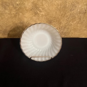 May include: A white, decorative bowl with a gold rim and a swirled pattern. The bowl is displayed on a gold-colored metal stand. The background is a textured, golden-brown color.
