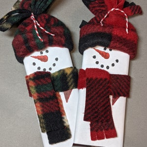 May include: Two snowman-shaped chocolate bars wrapped in silver foil. Each snowman has a red and black checked hat, a red nose, and black eyes. One snowman has a red and green checked scarf, and the other has a red and black checked scarf.