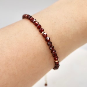 Pu&ograve; includere: Un delicato braccialetto con perline di granato rosso intenso sfaccettate. Le perline sono infilate con piccoli distanziatori color oro. Il braccialetto &egrave; mostrato su un polso, evidenziando il design e il colore del gioiello.